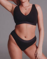A woman modeling a black two-piece lingerie set consisting of a V-neck bralette and matching high-waisted briefs, shown against a solid gray studio background.