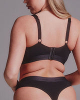 A woman shown from the back wearing a black bralette with shoulder straps and matching black underwear against a plain gray studio background.