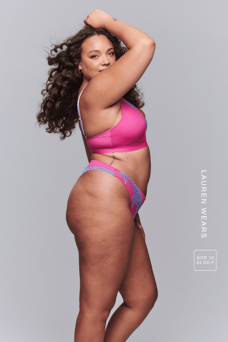 A woman standing with her side to and arms up wearing a pink and purple floral embroidered matching lingerie set 