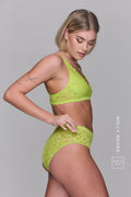 A woman standing with her side to, looking down wearing a lace lingerie set in the colour "Lime"