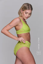 A woman standing with her side to, looking down wearing a lace lingerie set in the colour "Lime"
