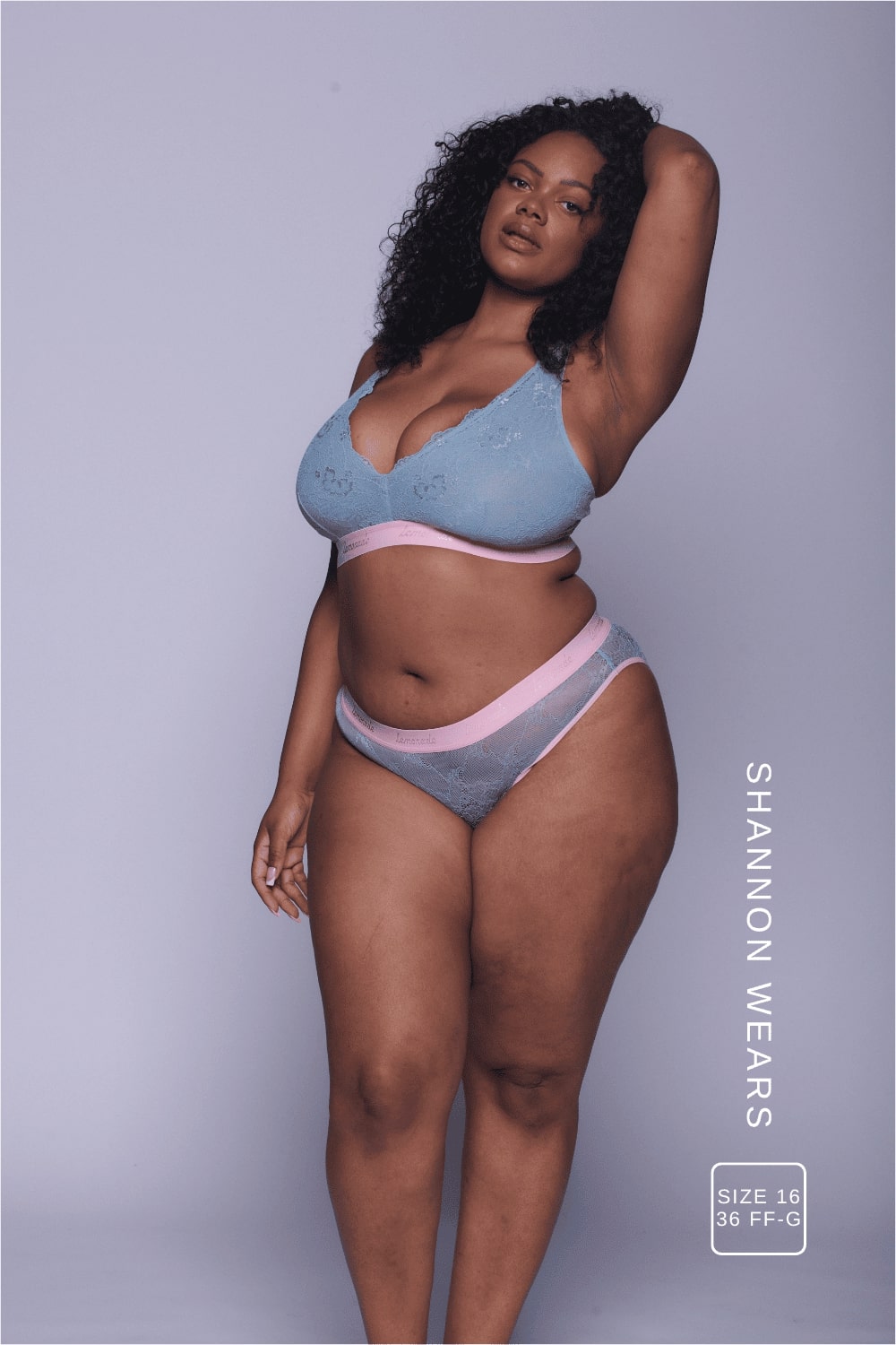 A woman standing with her hand in the hair wearing a light blue lace fuller cup bralette with a pink band with matching briefs 