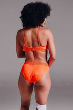 A woman standing with her back to wearing the signature lace bralette in neon orange paired with the matching brief 