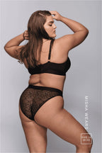 A woman standing with her back to and her arms up, wearing a black lace lingerie set including a black lace brief 