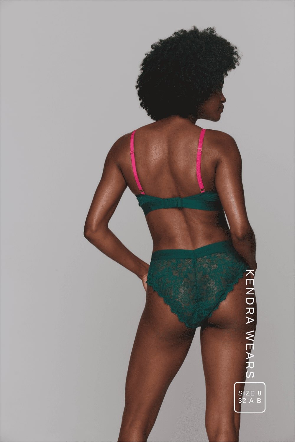 A woman standing with her back to modelling a dark green lingerie set with pink straps ^Kendra | Size: 32 A-B / 8